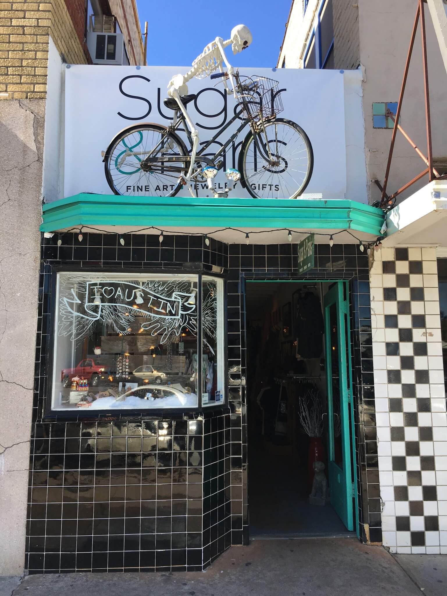 Exterior of Sugar & Paris store on South Congress, black tile with teal doorway and skeleton on bicycle over entrance